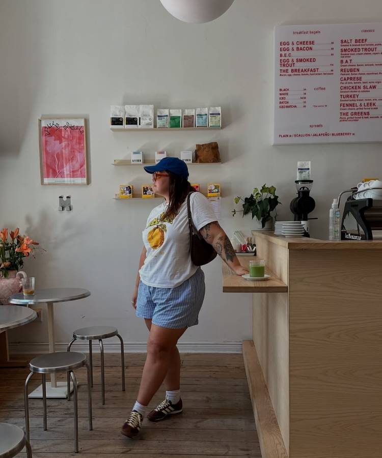 Uma mulher com um boné azul e óculos escuros, vestindo uma camiseta branca, **shorts verão 2026** azuis e brancos, meias brancas e tênis. Ela se inclina em um balcão de madeira em um café com um menu na parede.