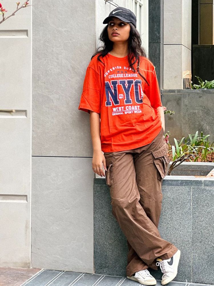 Mulher de pele clara usando look com camiseta oversized laranja, calça cargo marrom e boné. 