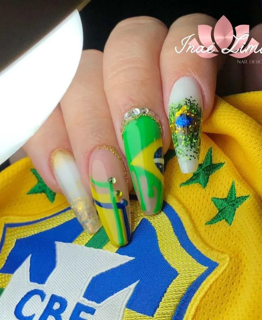 Imagem de mão com unhas decoradas para a Copa do Mundo 2022. Cores usadas, verde, amarelo, azul e branco. Indicador, branco com glitter nas cores da bandeira e com o formato dela. Médio, desenho de bandeira + pedras decorativas. Anelar, arte com as cores verde, amarelo e azul + pedras decorativas. Mínimo, branco com dourado. 