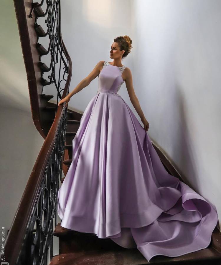 Uma noiva de cabelo preso, usando um elegante vestido de cetim lilás com saia volumosa e cauda, bordados nos ombros e cintura, posa em uma escadaria de madeira escura com corrimão de ferro forjado.







