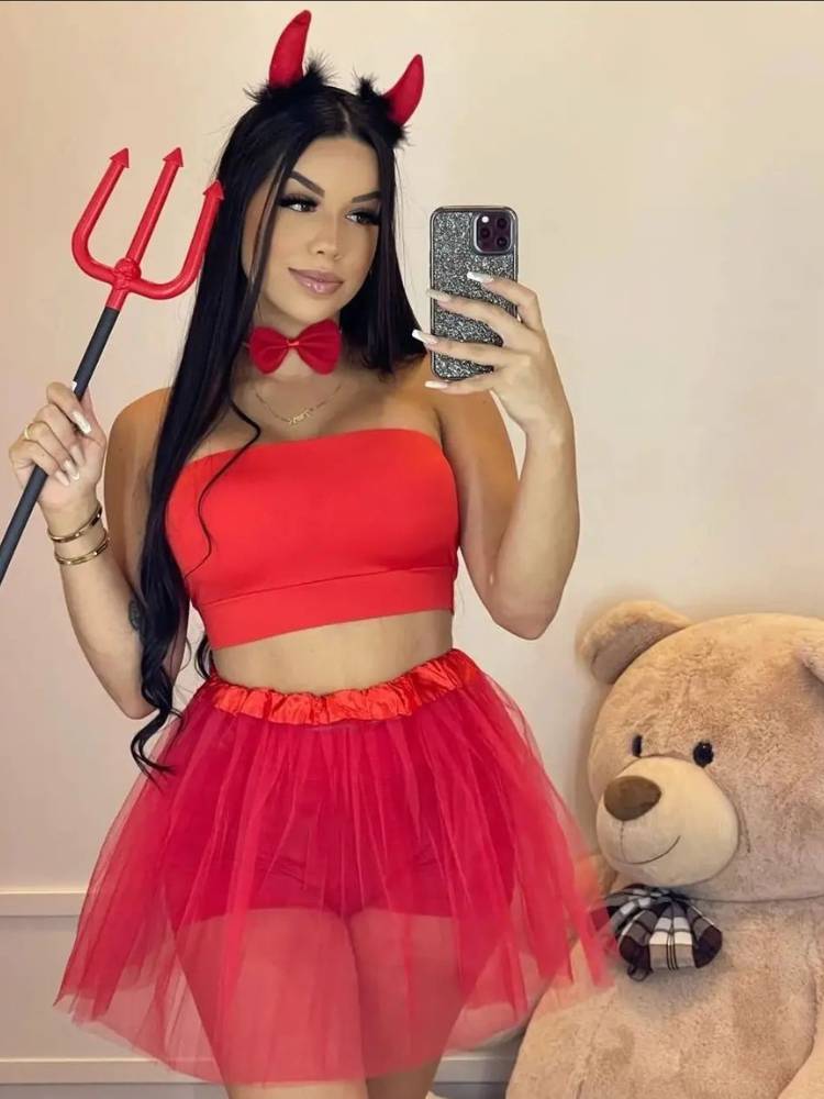 Mulher de pele clara e cabelo escuro com tridente vermelho, cropped vermelho, saia de tule e chifre de diabinha. 