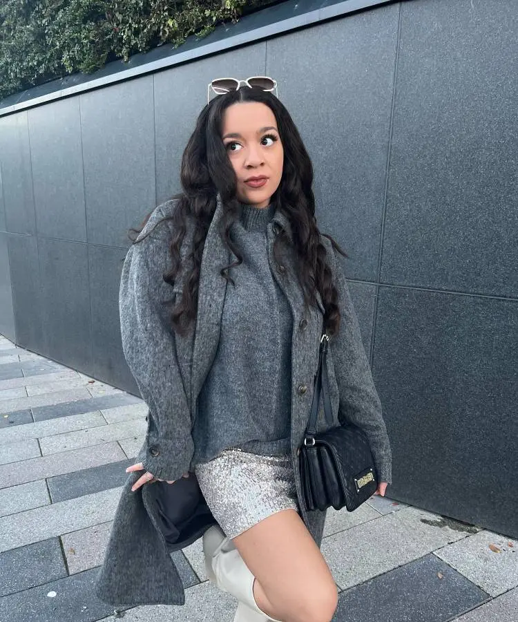 A mulher posa com elegância em um ambiente urbano, vestindo um conjunto cinza de casaco e suéter sobre uma saia prateada de paetês. Botas brancas, bolsa preta e óculos de sol no cabelo completam o visual sofisticado.