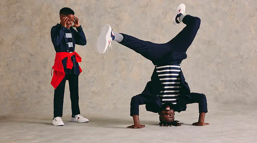 Filho de pele negra usando jaqueta jeans, calça preta e blusa branca e preta vendo o pai dançar usando as mesas roupas