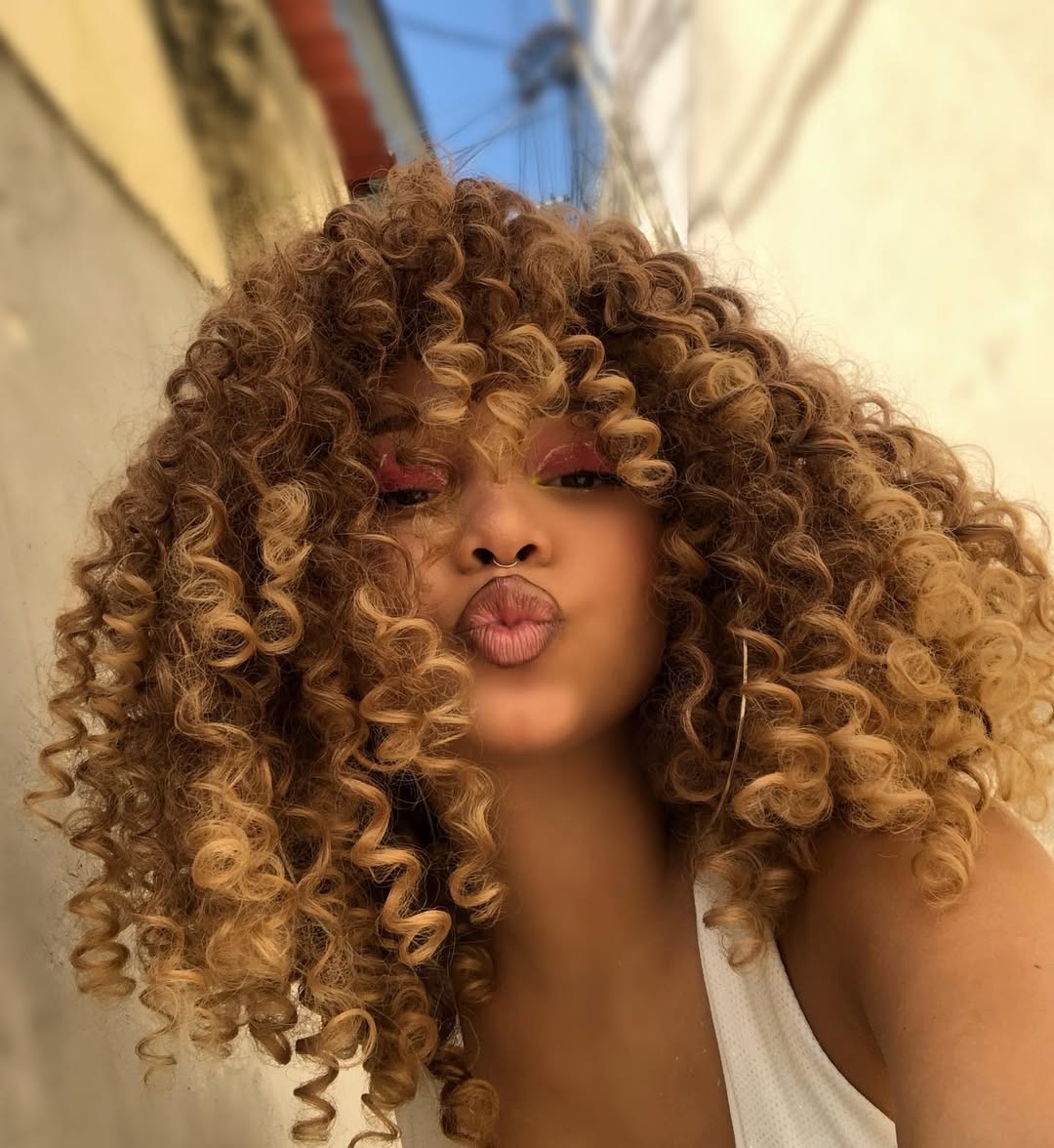 Mulher com cabelos cacheados volumosos em tons de castanho mel, vestindo uma regata branca. Ela usa maquiagem vibrante com sombras rosa e amarela, fazendo um gesto de beijo para a câmera, em um ambiente externo iluminado naturalmente.