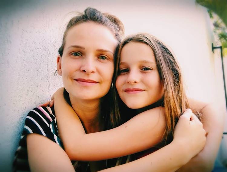 Uma mulher e uma menina sorriem para a câmera, abraçadas afetuosamente. A mulher veste listras e a menina tem cabelo longo. Elas estão encostadas em uma parede clara, transmitindo um momento de carinho e proximidade, irradiando felicidade e amor.
