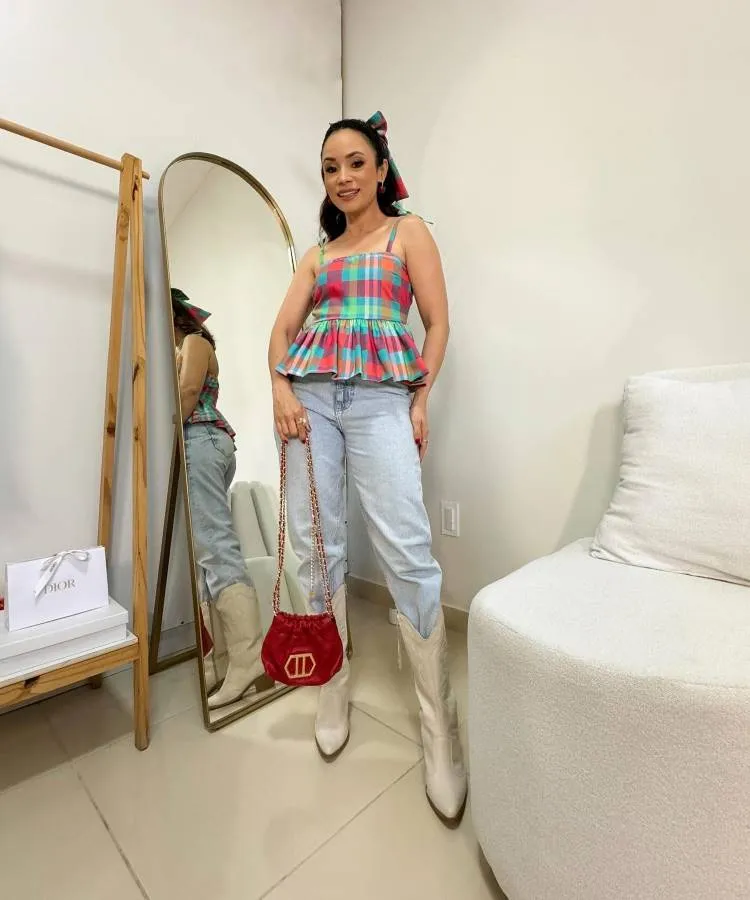 Mulher com blusa peplum xadrez colorida e jeans claros. Usa botas brancas estilo western e segura bolsa vermelha de veludo. Cabelo preso em rabo com laço. Está em ambiente claro, com espelho, cadeira branca e acessórios minimalistas ao redor.
