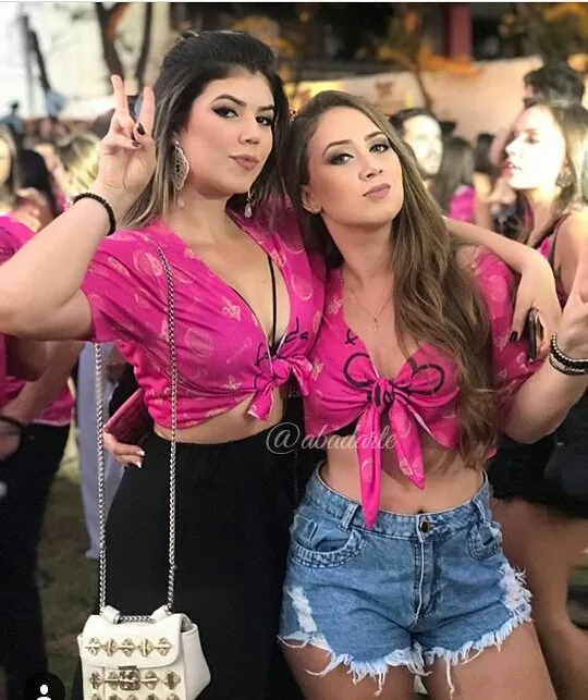 Duas mulheres de pele clara com abadá rosa customizado com nó, sendo que uma usa calça preta e outra usa shorts jeans