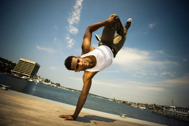 Rapaz negro fazendo passo de dança com o corpo usando uma só mão no chão, em local com vista para uma lagoa, com prédios e casas ao fundo