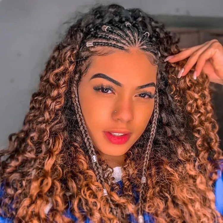 Mulher com trança nagô até metade da cabeça, com joias, seu cabelo é cacheado iluminado.