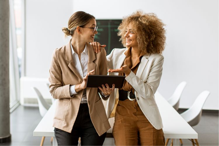 A imagem retrata duas mulheres empresárias engajadas em networking para mulheres. Uma mulher de cabelos claros e óculos segura um tablet, enquanto a outra, com cabelo afro, gesticula animadamente. Ambas sorriem, sugerindo uma colaboração bem-sucedida em um ambiente de trabalho moderno e luminoso.