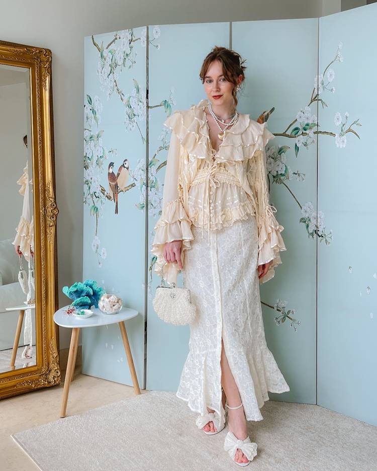 Mulher com look mermaidcore,  ela usa roupa clara saltos Claro e bolsa Clara com formato de concha ao fundo um painel com desenho de pássaros em uma árvore ao lado um espelho, uma mesa e ela está em cima de um tapete
