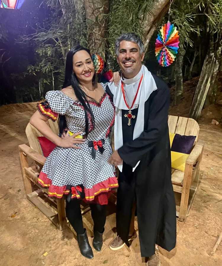 Casal com roupa junina, ela de vestido de poá com laço da Minnie e ele de padre da tradicional quadrilha