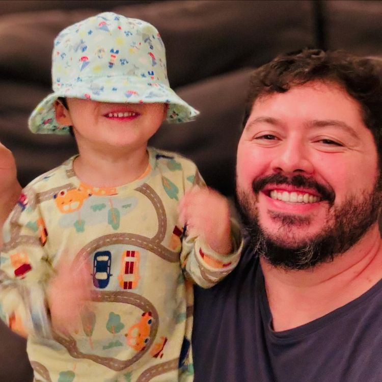 Atila de blusa preta sorrindo ao lado de criança usando roupa colorida e bucket hat cobrindo o rosto