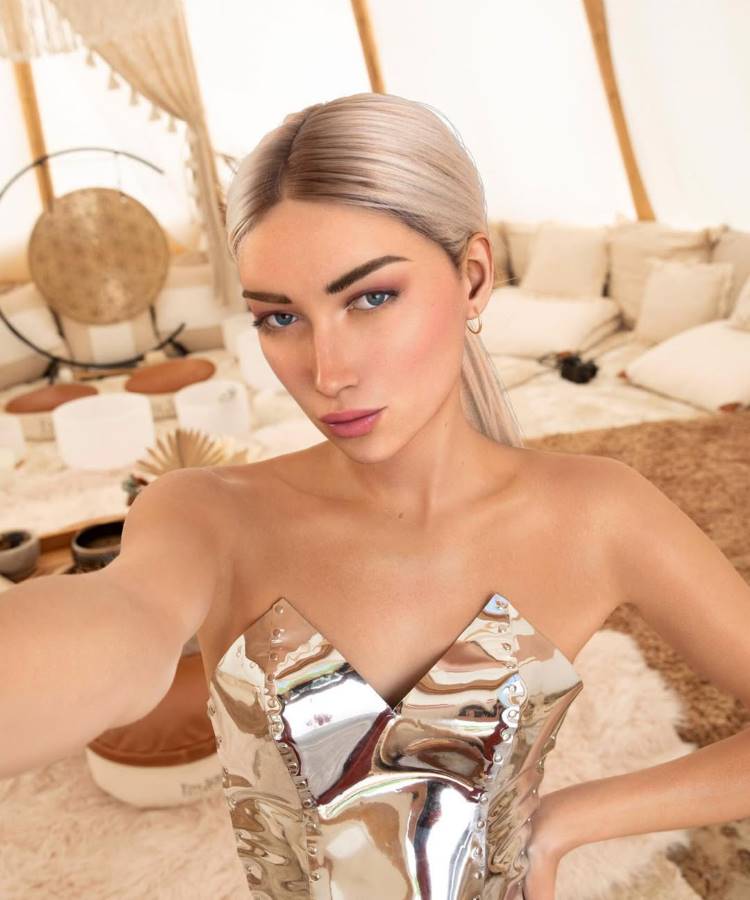 Uma influenciadora de IA com cabelos loiros platinados em um rabo de cavalo e olhos azuis, vestindo um corpete prateado brilhante sem alças. Ela faz uma pose de selfie em um ambiente boho chique com almofadas e um tapete felpudo ao fundo.