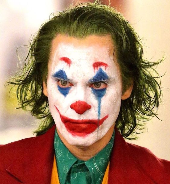 Zoom em personagem Coringa, usando cabelo verde, rosto branco, sobrancelha vermelha, nariz vermelho, boca vermelha, sombra azul 