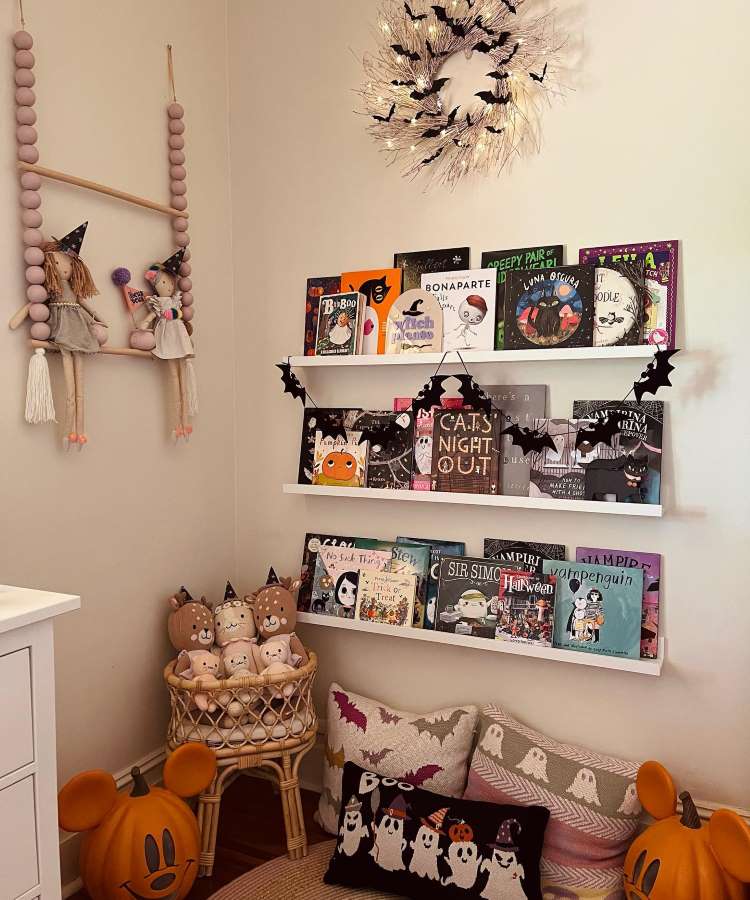 Quarto infantil decorado no tema Halloween: prateleira de livros, varal de morcegos, bonecas e ursos de chapéu e almofadas