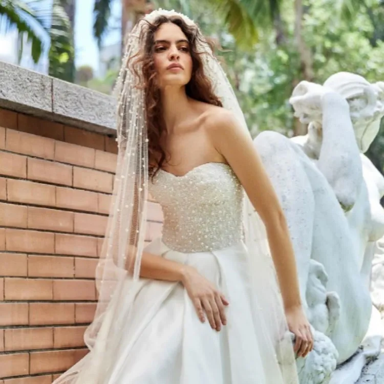 Mulher de pele clara ao lado de muro com tijolos à vista, usando vestido de noida com corset bordado de pérolas, saia evasê lisa e véu com pérolas