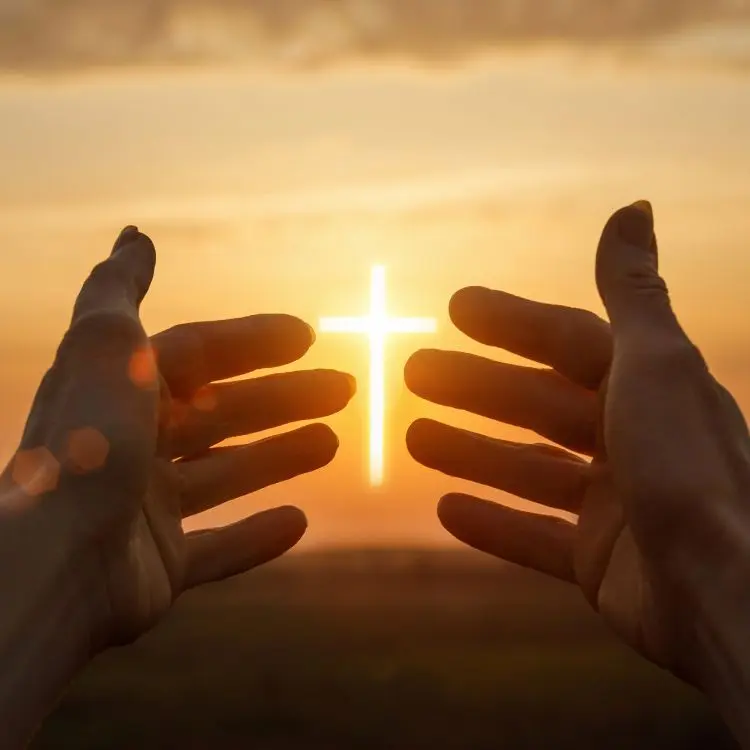 Mãos abertas em direção a uma cruz iluminada pelo sol, simbolizando fé e oração. O pôr do sol ao fundo irradia luz quente e dourada, criando uma atmosfera espiritual e contemplativa em meio à paisagem natural desfocada.