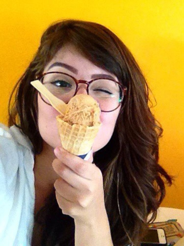 Selfie de Thais em 2015, fundo amarelo, óculos, cabelo longo e sorvete na frente do rosto 
