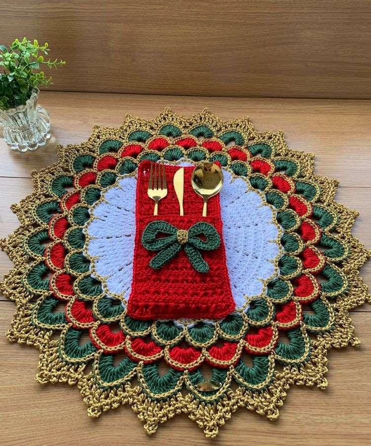 Sousplat de crochê colorido com detalhes em vermelho, verde e dourado. Porta-talheres vermelho com laço verde segura talheres dourados. Ao lado, um vaso de vidro com planta sobre mesa de madeira.