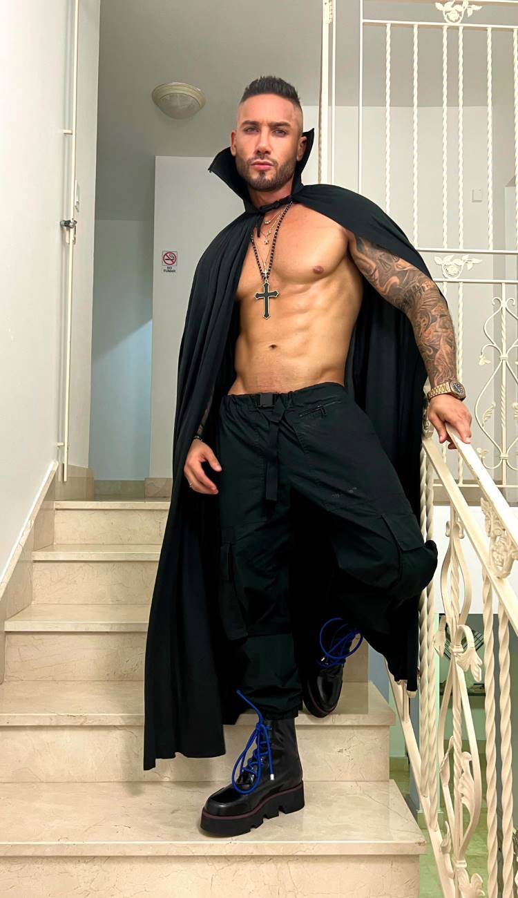A imagem mostra um homem em pé em uma escada, vestindo uma capa preta que revela seu tronco musculoso. Ele usa calças pretas e botas com cadarços azuis. Um colar com um crucifixo pendurado destaca-se em seu peito. O ambiente é iluminado, com paredes brancas e um corrimão de metal ao fundo.


