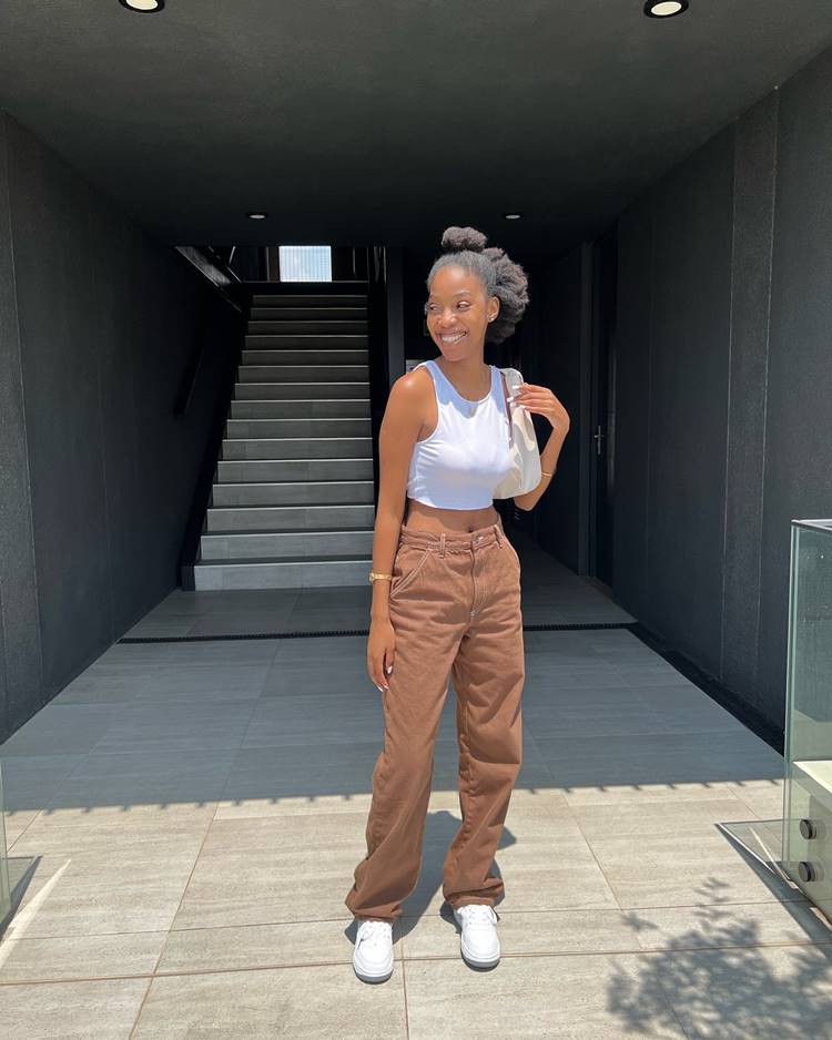 Garota negra usando regata cropped branca, calça marrom, tênis branco e bolsa branca, em local com escada.