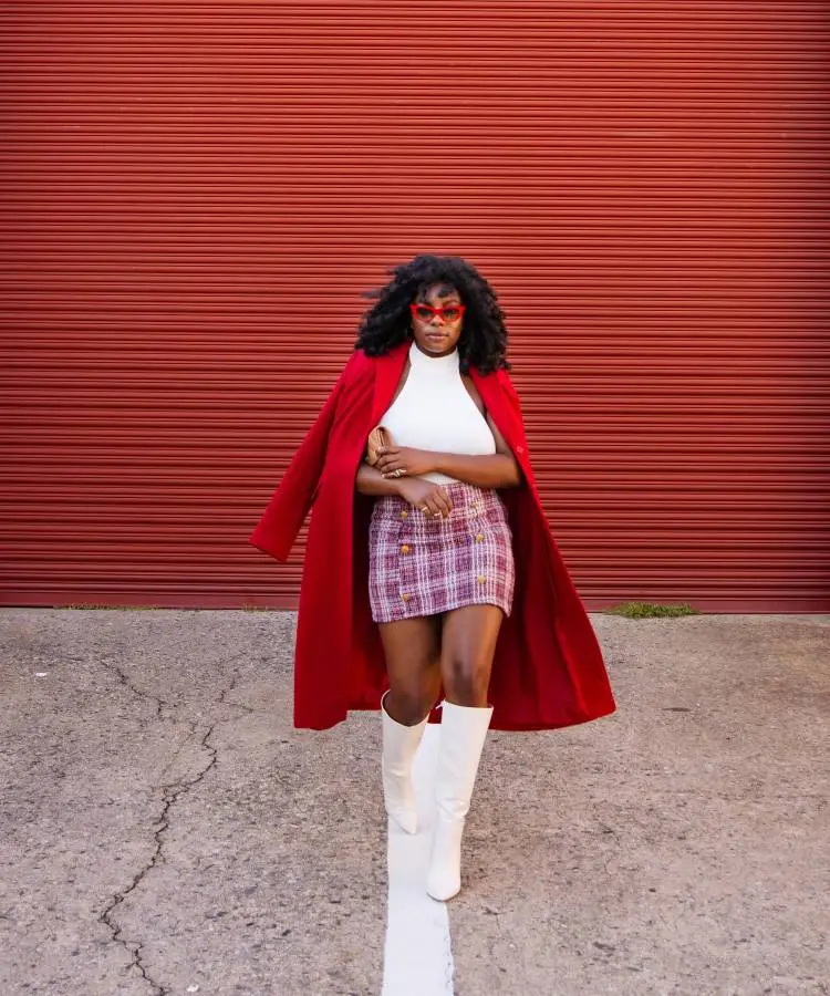 Uma mulher com cabelo encaracolado e óculos de sol vermelhos em forma de coração usa um sobretudo vermelho vibrante sobre uma blusa de gola alta branca e uma minissaia xadrez em tons de roxo e rosa. Ela completa o look com botas brancas de cano alto.