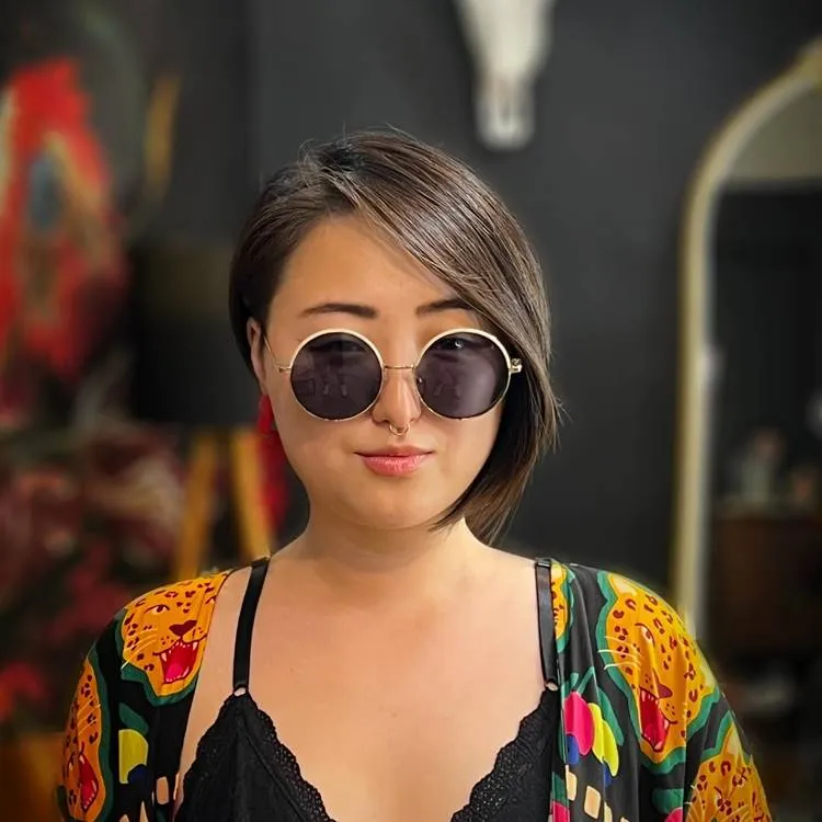 Mulher oriental usando óculos de sol, e roupa com estampa de onça em fundo de salão de beleza desfocado. Seu rosto é redondo e seu cabelo é um corte curto com franja lateral longa