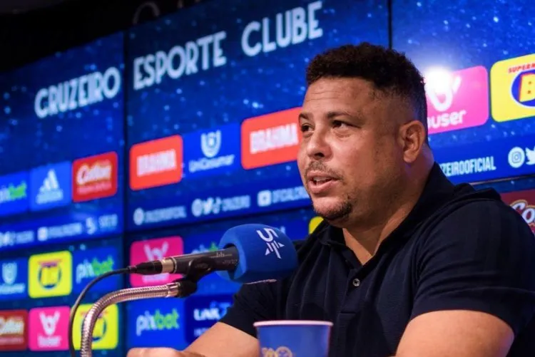 A imagem mostra um homem sentado em coletiva de imprensa, falando ao microfone azul com o logotipo "Cruzeiro". Usa camisa preta, cabelo curto e exibe expressão neutra. Ao fundo, painéis com logotipos e o nome "Cruzeiro Esporte Clube".