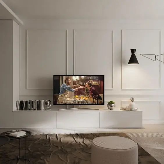 Sala com paredes brancas com moldura, luminária preta na parede, aparador branco com decoração, tapete bege, puff bege e mesas de centro ao canto