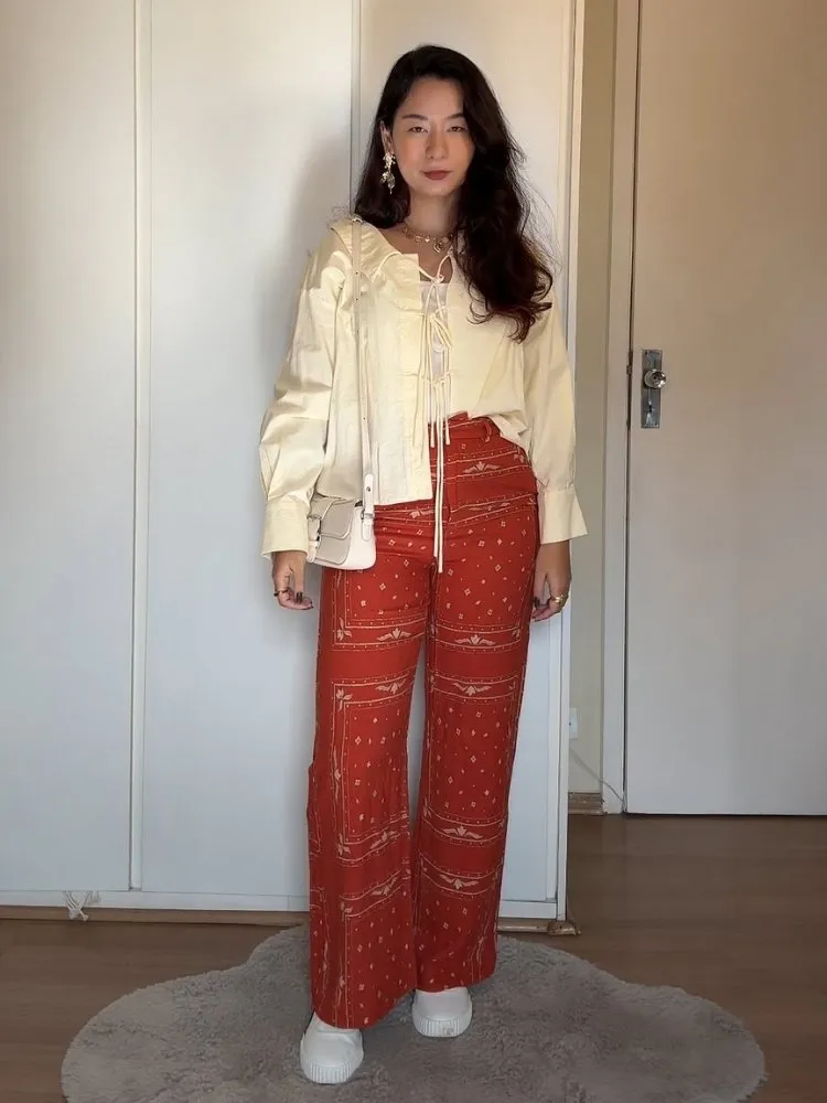 Mulher de origem asiática usando camisa amarelo-manteiga com amarração frontal, calça vermelha estampada e tênis branco 