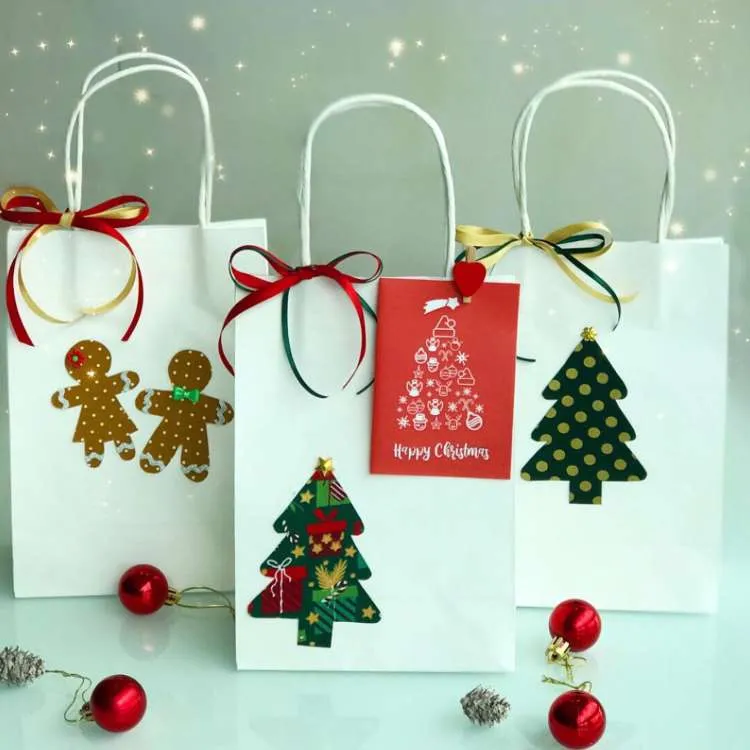 Três sacolas brancas de papel enfeitadas com itens natalinos, como árvores e biscoitos.