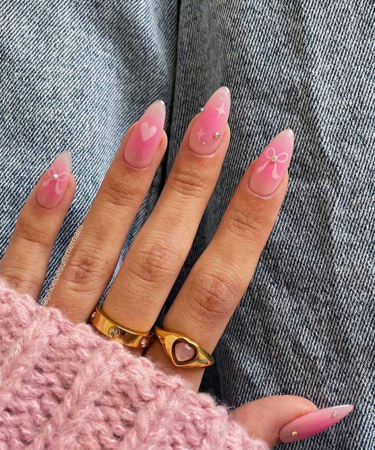 A imagem destaca unhas decoradas em tons de rosa com desenhos delicados de corações, laços e estrelas brancas, além de pequenos cristais. A mão exibe anéis dourados e está sobre um jeans, complementada por um suéter rosa macio.