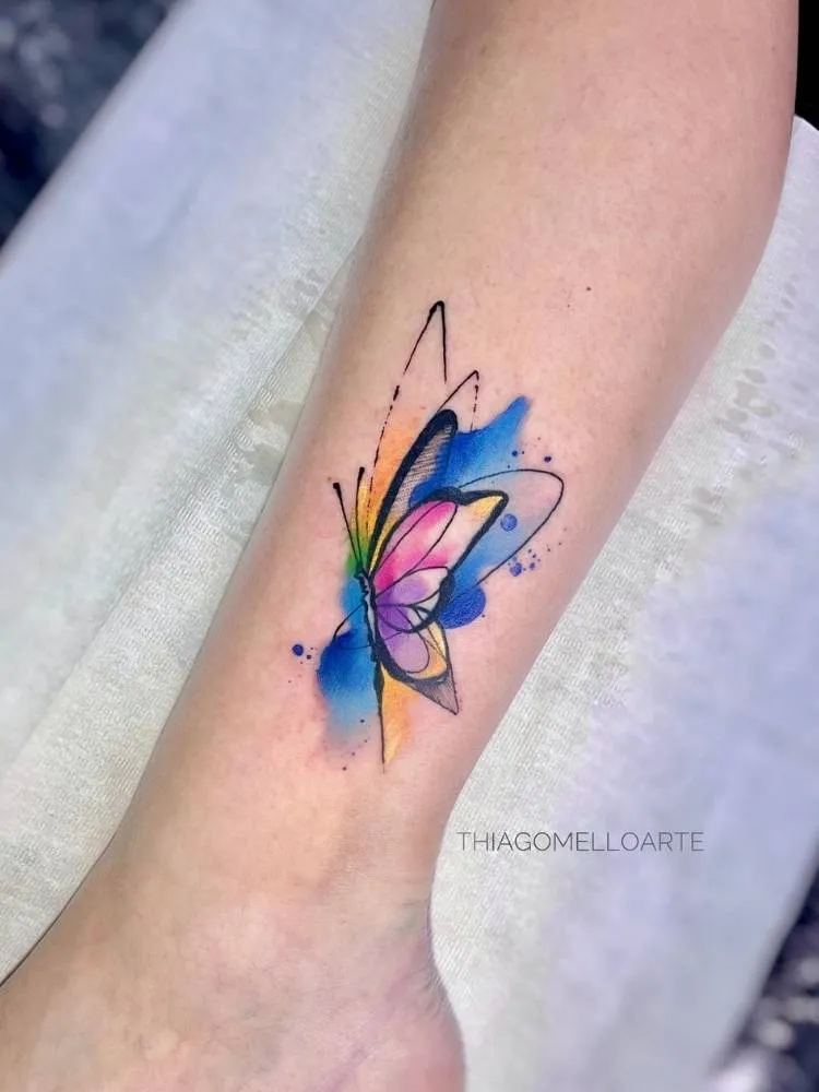 Tatuagem de borboleta com traços finos e grossos e desenho um pouco mais minimalista, mas com efeito aquarela nas cores azul, rosa e amarelo. Foi feita no braço de uma mulher de pele clara. 