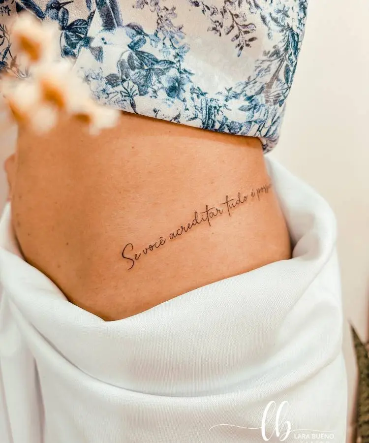 A imagem mostra uma tatuagem delicada na lateral do corpo de uma pessoa, com a frase "Se você acreditar tudo é possível." A pele está parcialmente coberta por uma blusa com estampa floral azul e branca. A cena é iluminada suavemente, destacando a simplicidade e elegância da tatuagem.


