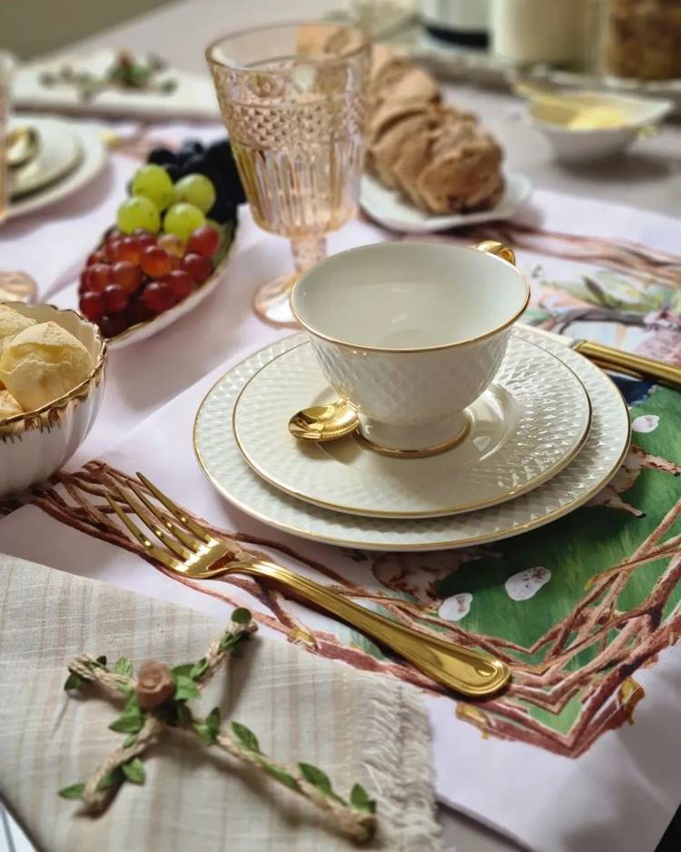 Uma mesa de café da manhã decorada para a Páscoa Cristã: conjunto de taças de cerâmica com detalhes dourados, talheres dourados, alimentos como pão, frutas, uva, pão de queijo, manteiga, taças de vidro, guardanapo xadrez, cruz sobre o guardanapo e sousplat com imagem de pintura de paisagem com Cordeiro.