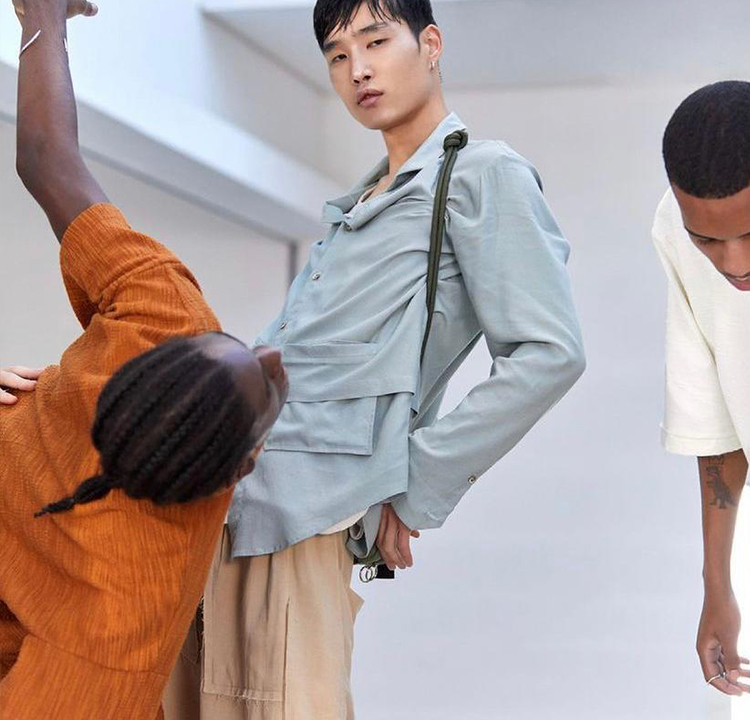 Três modelos, um usando uma blusa laranja, um usando uma blusa azul de botão e uma calça bege, o outro está quase fora do enquadramento da foto.