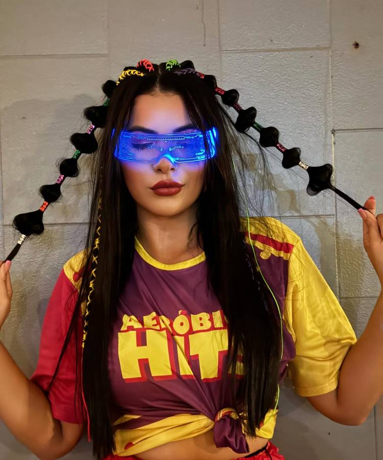 A imagem mostra uma mulher com cabelo longo estilizado em tranças decoradas com elásticos coloridos e bolhas pretas. Ela usa óculos futuristas com luz azul brilhante e veste uma camiseta vibrante escrita "Aeróbica HITs".