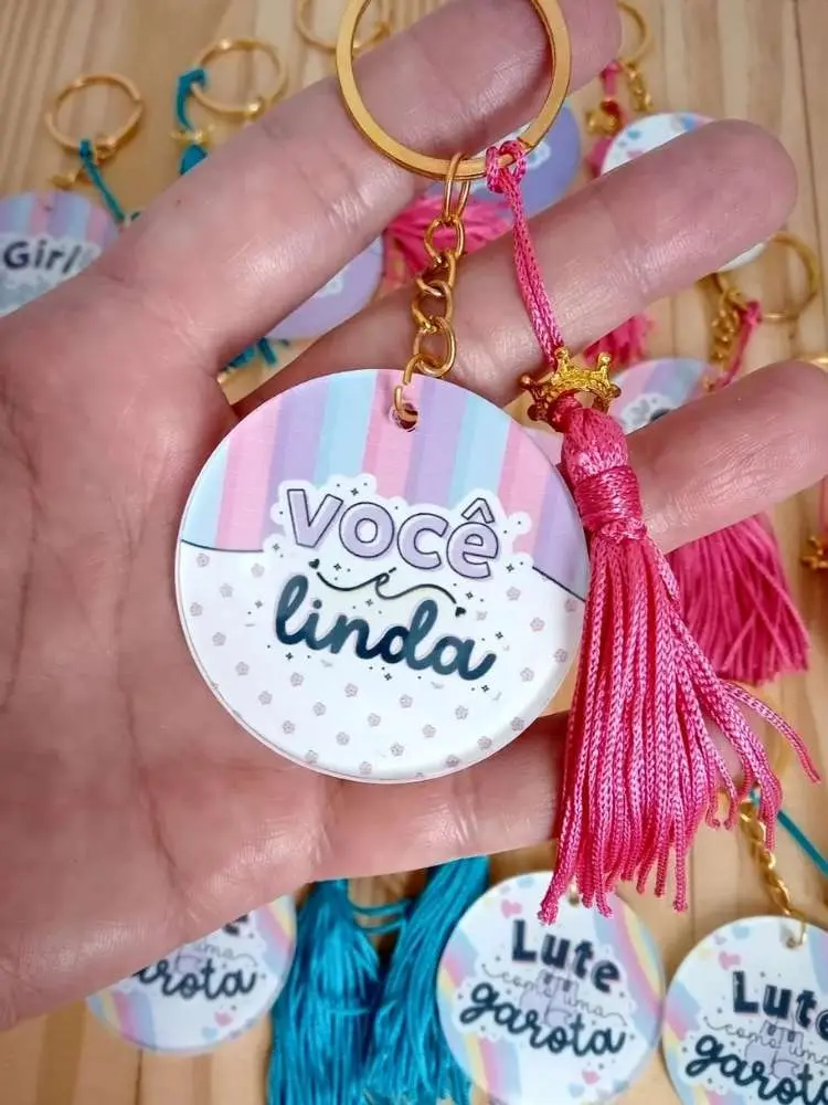 Chaveiro redondo com detalhe em rosa escrito "você é linda" e outros exemplos ao fundo