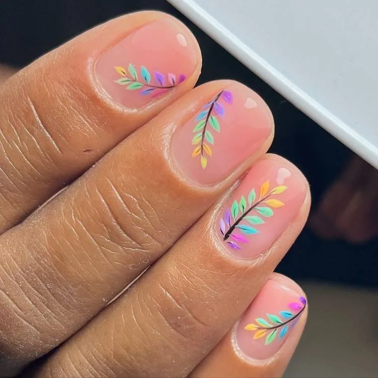 A imagem mostra um close-up de uma mão exibindo quatro unhas com manicure. As unhas estão pintadas com um esmalte de cor nude translúcido, permitindo que a cor natural das unhas apareça. Em cada unha, há um design intrincado de pequenas folhas em um padrão simétrico que se estende da base até a ponta. As folhas são coloridas em vários tons, incluindo roxo, verde, azul e amarelo, criando uma aparência delicada e colorida contra o fundo neutro do esmalte.

