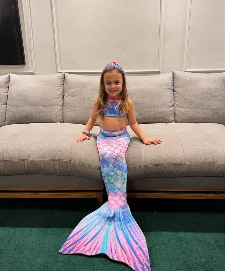 Menina fantasiada de sereia, sentada em um sofá cinza. Ela usa uma cauda azul e rosa com escamas brilhantes, um top combinando e uma tiara decorada. O ambiente possui parede branca, sofá acolhedor e carpete verde escuro.





