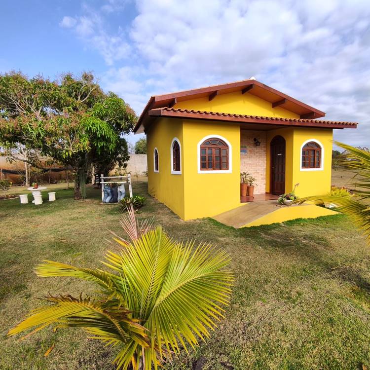 Imagem de fachada de casa simples e pequena na cor amarela. ELa está em um jardim gramado, com pé de manga e uma palmeira pequena. Há uma mesa branca abaixo do pé de manga. A entrada da casa possui uma rampa simples, que dá acesso à porta principal