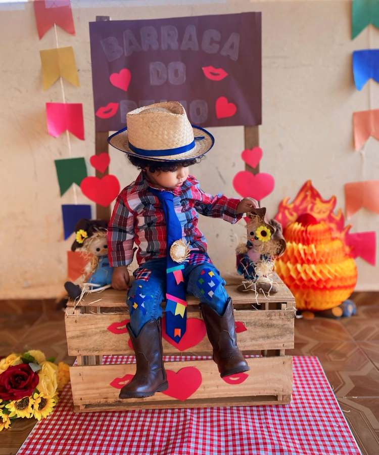 Menino usando camisa xadrez, calça decorada com retalhos, gravata decorada, bota de cano longo e chapéu de palha para festa junina.
