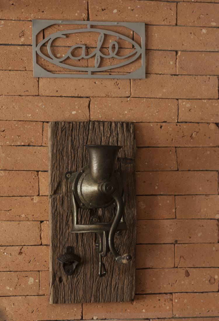 Na parede de tijolinhos de uma cozinha rústica moderna, um moedor de café antigo de ferro fundido está montado em uma tábua de madeira rústica, com um abridor de garrafas ao lado. Acima, um letreiro "Café" de metal.







