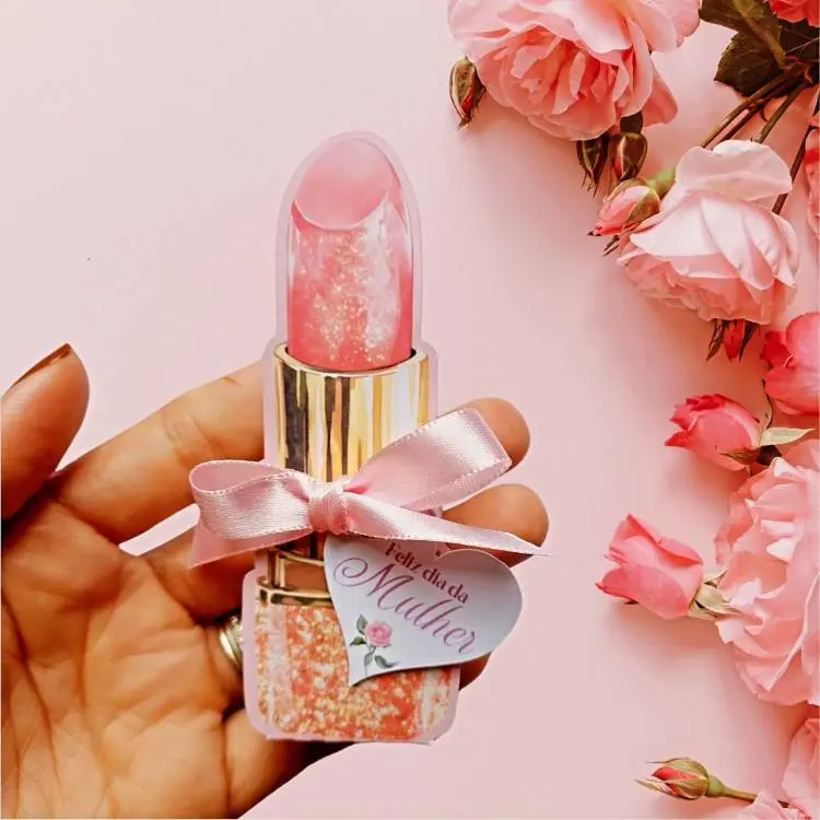 Lembrancinha em formato de batom com detalhes brilhantes, laço rosa e tag delicada escrita "Feliz Dia da Mulher". Fundo decorativo em tom rosa com flores delicadas, destacando-se como um presente criativo e especial para homenagens femininas.