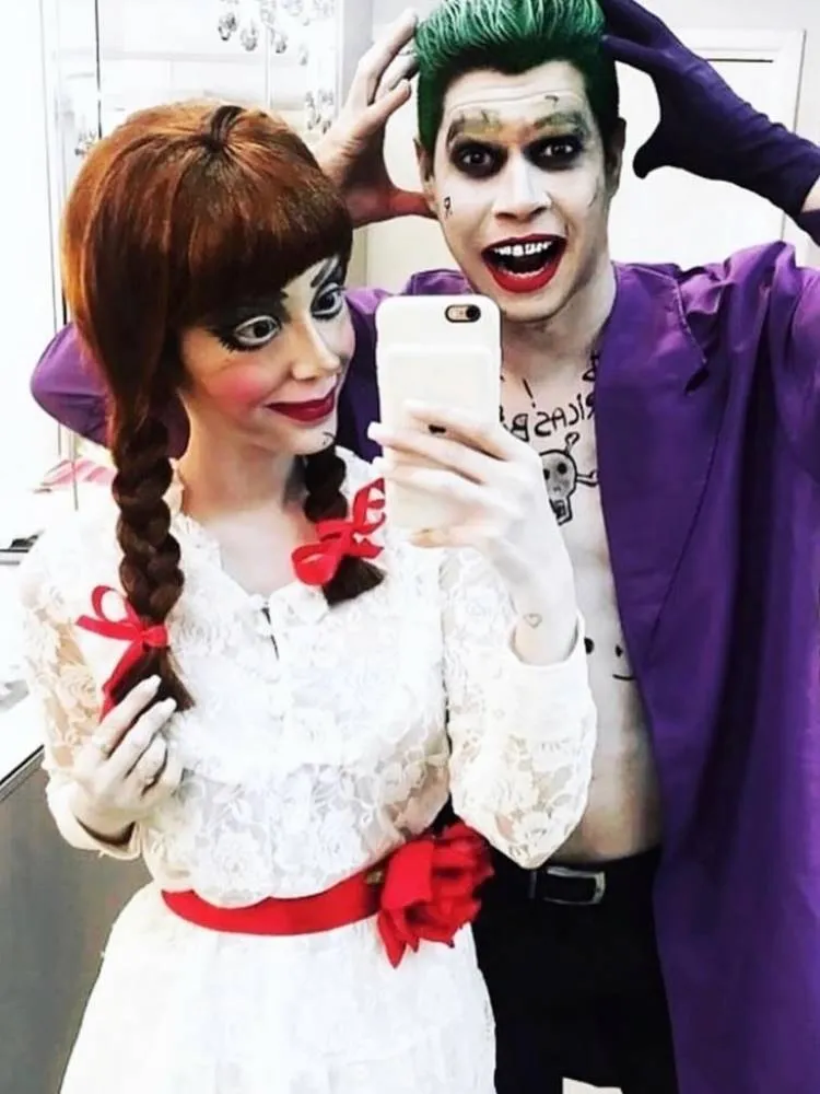 Casal de influenciadores Rica de Marré e Adrão Meron fantasiados de Anabbelle e Coringa para o Halloween. Ela usa maquiagem da boneca, tranças com laço vermelho e vestido branco. Já ele usa maquiagem pálica, cabelo verde, desenho de tatuagens e terno roxo 