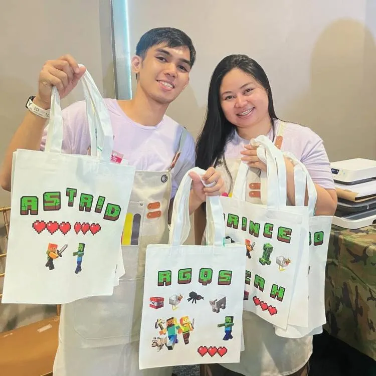 Um casal sorridente segura sacolas de tecido brancas e personalizadas, com o nome "ASTRID" em um e "PRINCE" no outro, ao lado de personagens de jogos como Minecraft, com corações e elementos do jogo.

