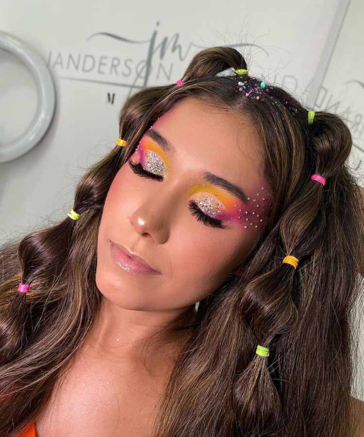 Mulher de pele clara com maquiagem amarela e rosa com glitter e strass. Ela também usa elásticos coloridos no cabelo 