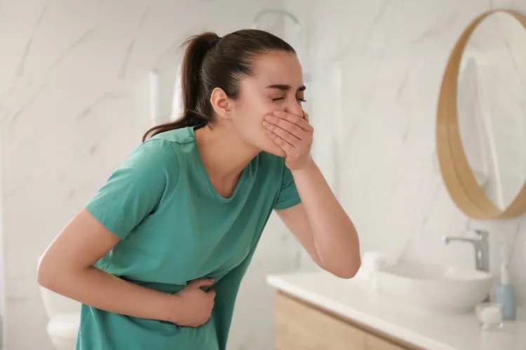 Mulher de pele clara e cabelo escuro usando camiseta verde e com a mão na barriga e outra tampando a boca, como se estivesse com enjoo