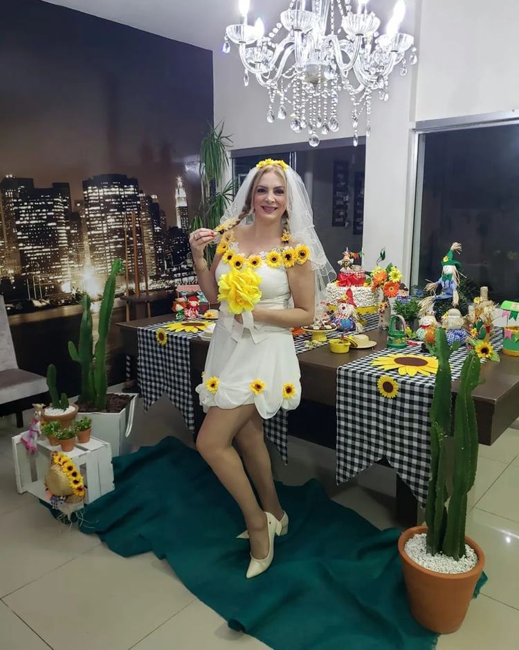 Noiva de festa junina com vestido decoração com flores de girassol em frente a uma mesa decorada com objetos temáticos e comidas típicas